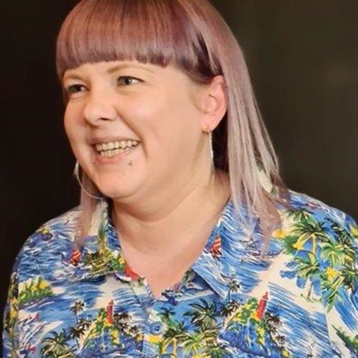 Claire has sleek, light purple hair and is wearing a vibrant blue patterned blouse.