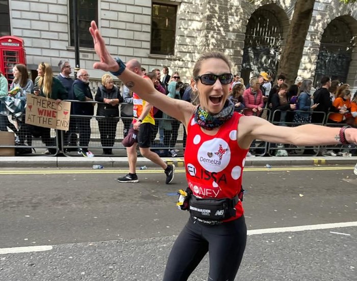A woman wearing a Demelza running vest, holds her hands in the air, smiling as she runs in the London Marathon.