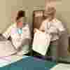 Two volunteers in white uniforms smile while changing bed linens on a hospice bed.