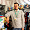 A young man with black hair leaning on a shelf at a Demelza charity shop smiling at the camera