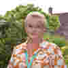 Demelza CEO Lavinia Jarrett stands in the hospice gardens, wearing a bright top and jewellery, smiling warmly.