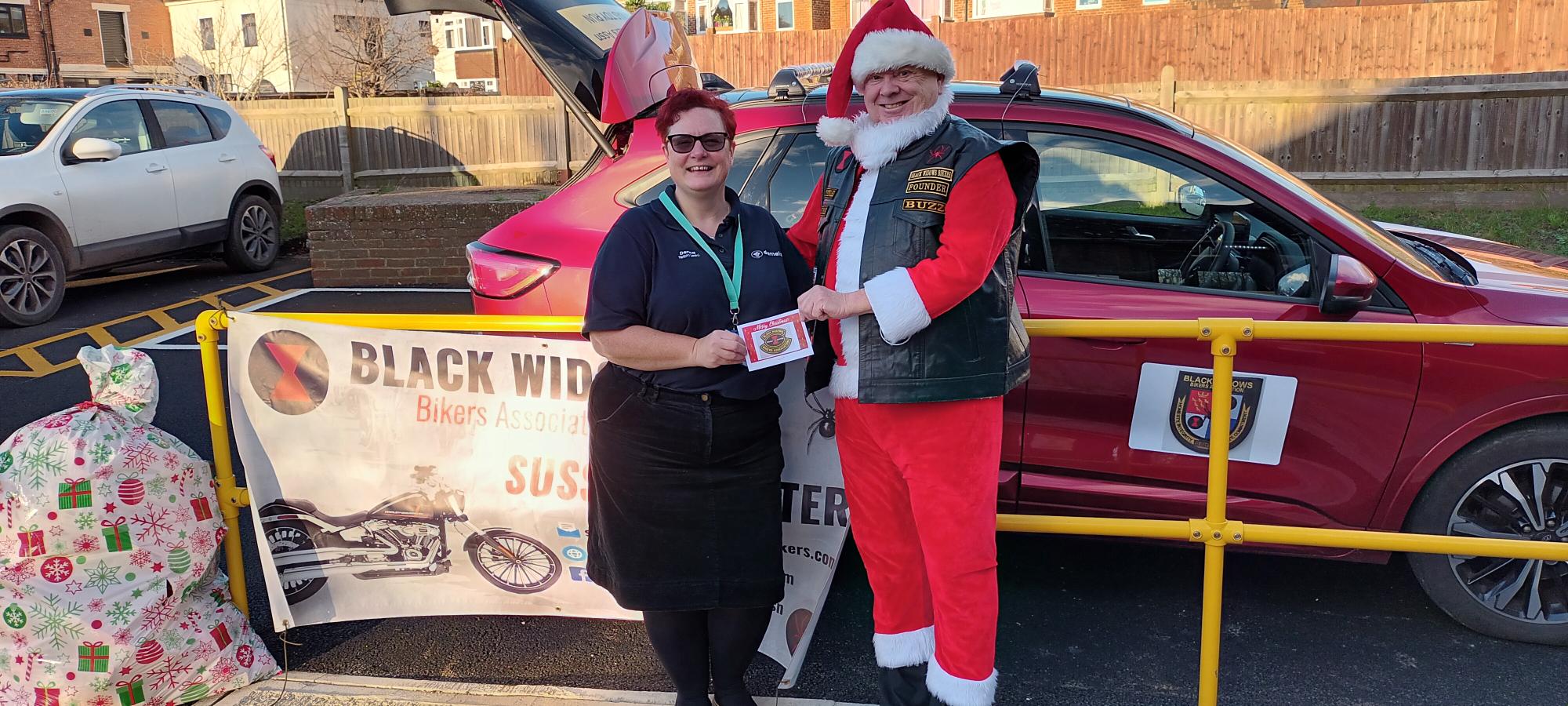 Two people stand by a red car with a 'BLACK WIDOW Bikers Association SUSSEX CHAPTER' banner. One is dressed as Santa; the other holds a card. Gift bags are on the left.