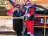 Two people stand by a red car with a 'BLACK WIDOW Bikers Association SUSSEX CHAPTER' banner. One is dressed as Santa; the other holds a card. Gift bags are on the left.