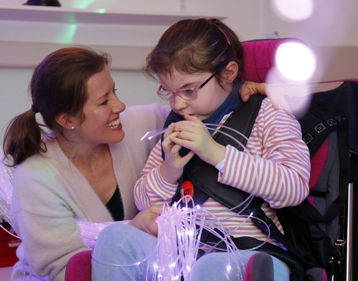 A young girl sits in her wheelchair, with mum crouched besides her smiling. They are in playing with glow sticks in the sensory room.