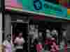 Three people outside a Demelza charity shop with a colorful sign and window display.