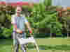 Man in white t-shirt with "demelza" wording pushing an orange and white STIHL lawnmower on a grassy lawn, with trees and bushes in the background.