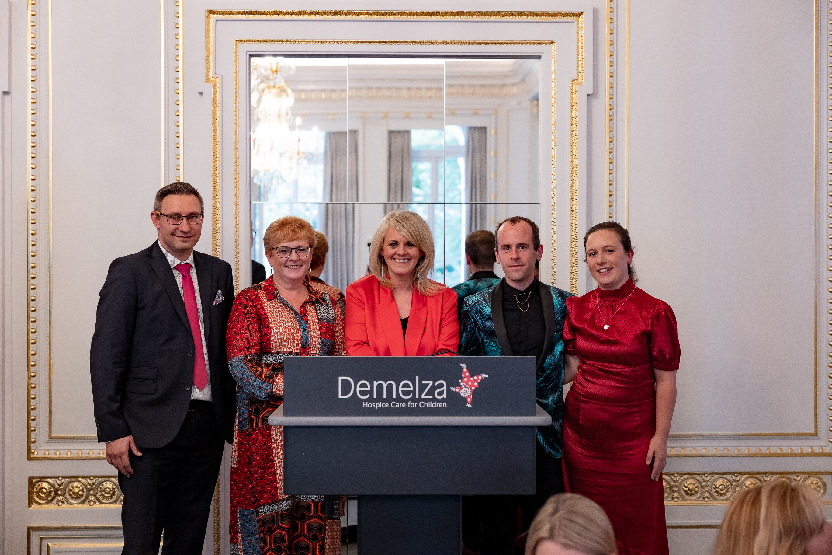 Five smartly dress people, stand being a Demelza branded lectern at the autumn lunch.
