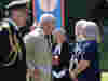 Group gathered outdoors near a blue and red banner with emblem. Includes man in military uniform and King Charles III in a beige suit.