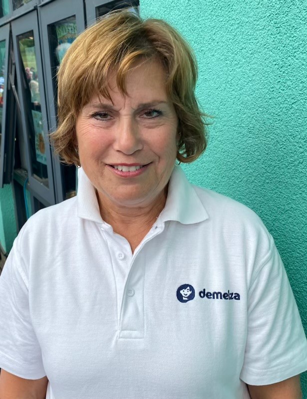 Person wearing a white polo shirt with "demelza" logo, standing in front of a turquoise wall with windows.