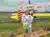Young person in white Demelza t-shirt and blue shorts holding a plush toy, standing in front of a yellow biplane with red and yellow striped wings.