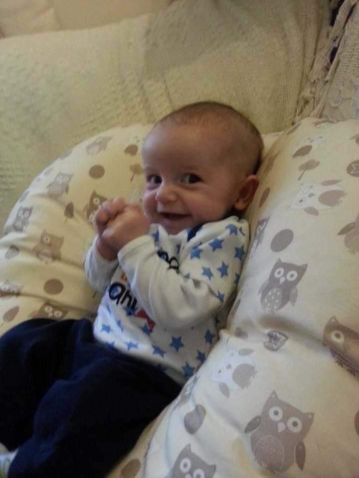 Baby Ronnie sits on an owl print cushion, smiling and holding his hands together.