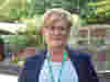 Woman stands outdoors wearing blue blazer, light blue top, and green Demelza lanyard. Background includes greenery and structures.