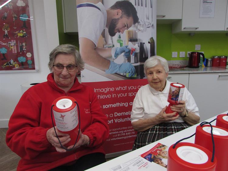 The ladies are sitting behind a fundraising desk, holding red Demelza collection pots.