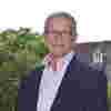Trustee Richard Finn stands proudly in sunny gardens, wearing glasses, a pink shirt and navy blue blazer.