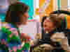 A person with short hair wearing a colourful, patterned sweater interacts with a child seated in a specialized chair. The child, dressed in a white shirt and green trousers, faces the person. The background features various colours and objects, in an indoor setting therapy room.