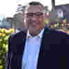 Trustee John Simpson smiles in sunny gardens, wearing a white shirt, smart blue jacket and black framed glasses.