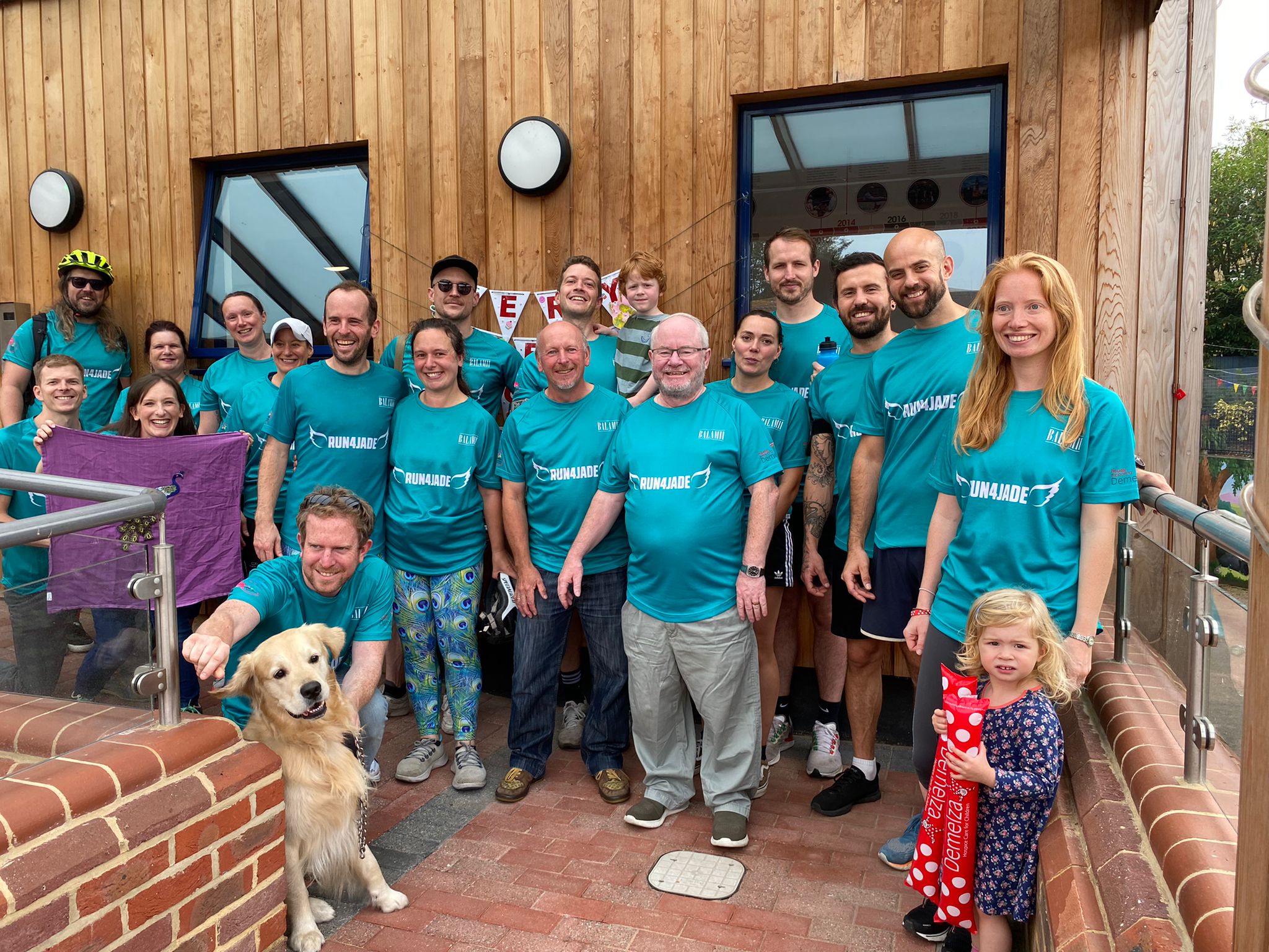 A team of fundraisers stand together outside Demelza's Eltham hospice, wearing matching teal 'Running 4 Jade' t-shirts.