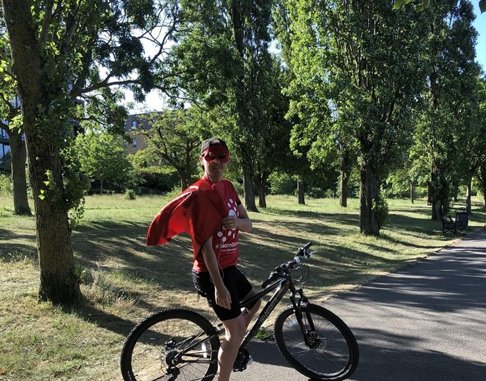 A man wearing a Demelza t-shirt, red cape and red superhero mask, riding his bike in a park.