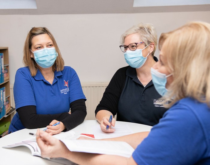 Three nurses, wearing blue polo tops and face coverings, sit around a round table, discussing paperwork.
