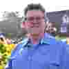 Trustee Dr Karl Holliman smiles in sunny gardens, wearing a blue checked long-sleeved shirt and black framed glasses.