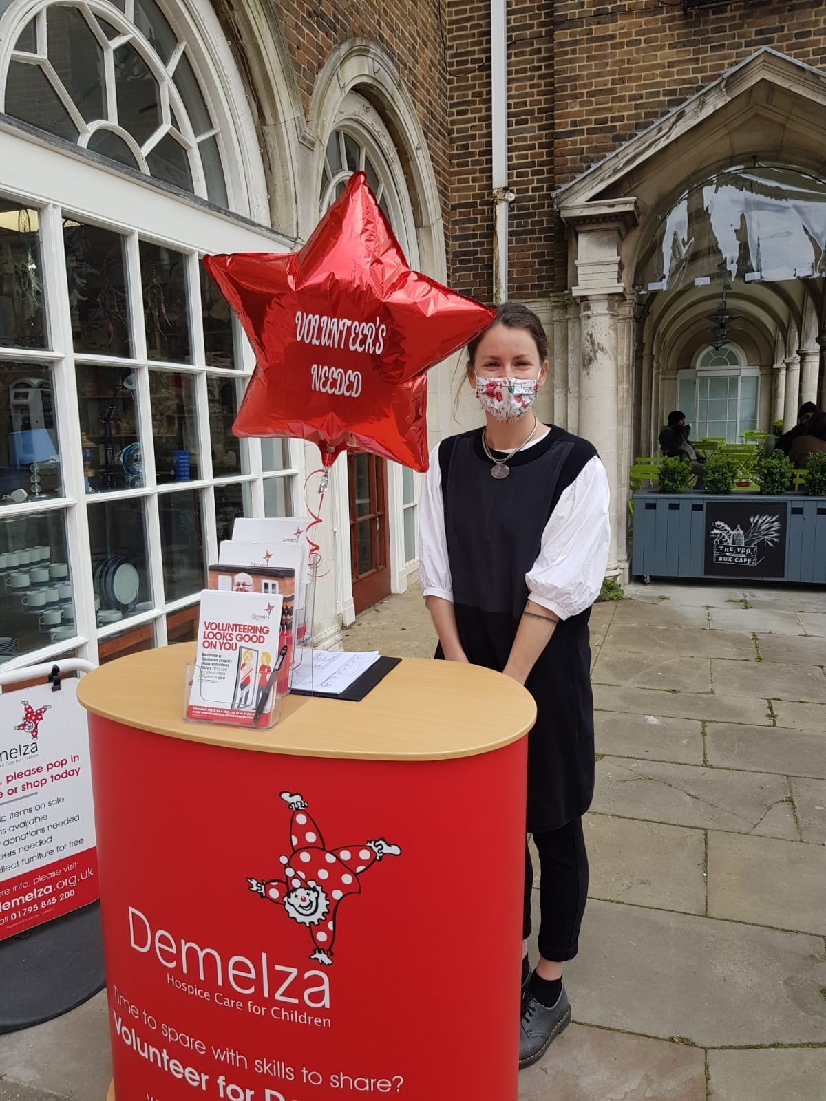 A woman wearing a face mask, standing behind a volunteer recruitment stand, outside the Canterbury charity shop.