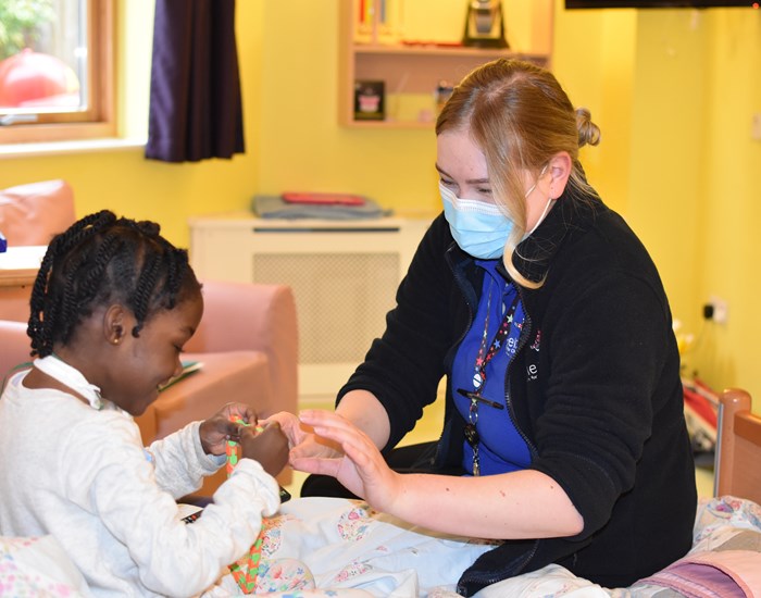 A nurse, wearing a medical face mask, plays with a young girl who is sitting on her bed.