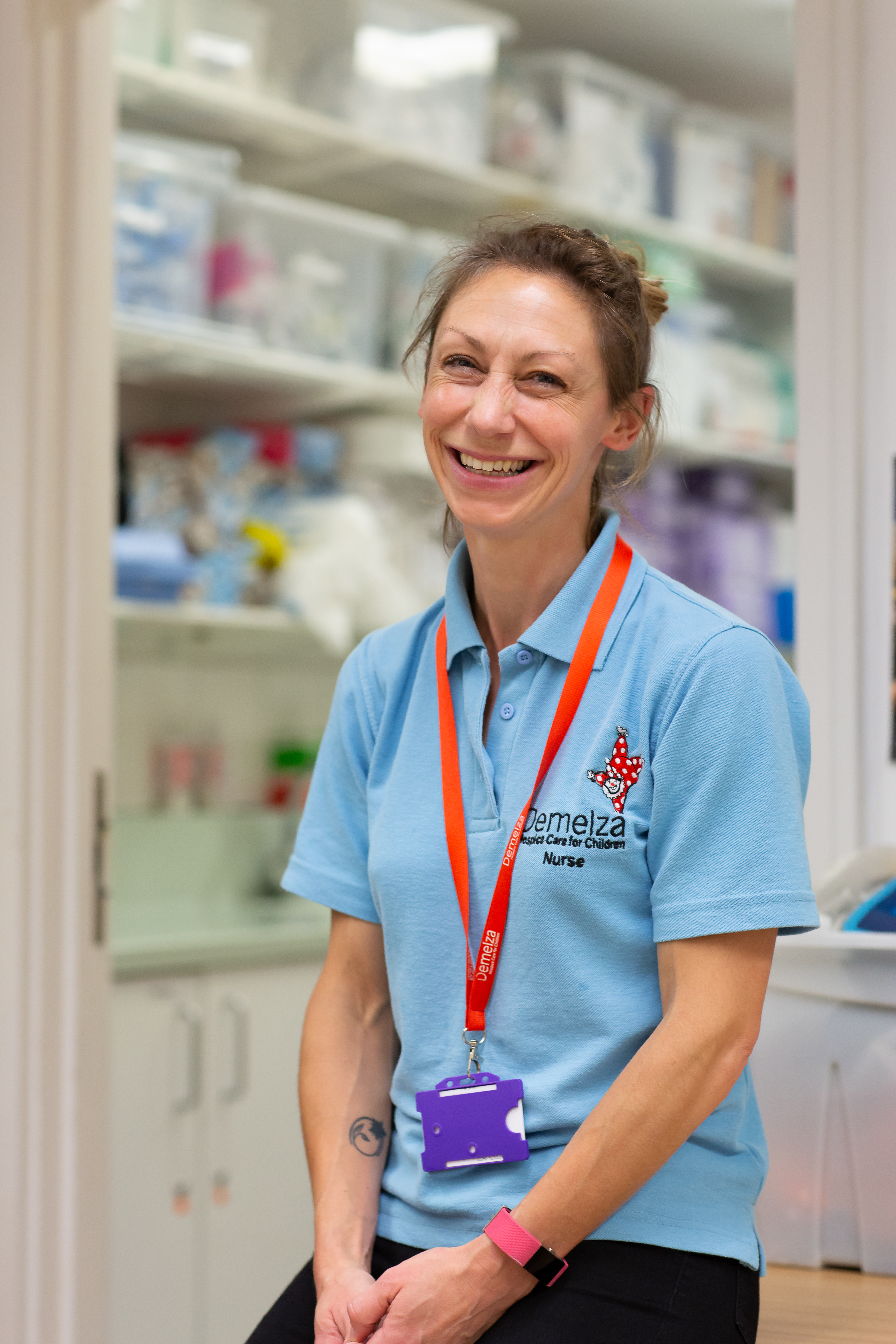 Demelza's Senior Nurse, Melissa, sits perched on a desk, smiling and wearing a light blue polo top.
