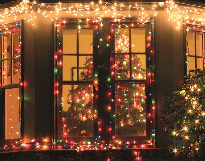 The front windows of a house are decorated with Christmas lights and a Christmas tree sits inside, with sparkling lights.