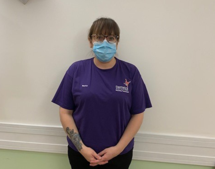 Becky is wearing her new, purple, Nursing Associate uniform, standing against a white wall.