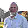 Chair of ACG Alex Parry-Jones stands smiling in sunny gardens, wearing a white and green checked shirt.