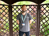 Man stands in front of wooden lattice, wearing grey polo and Demelza ID badge. Background includes greenery and plants.