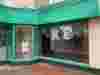 Storefront with green trim showing white Demelza t-shirts on a mannequin and inside; a colorful "VOLUNTEERS NEEDED" banner is displayed.