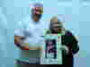 A man in a white shirt and a woman in a black shirt holding up a photo of their son who sadly passed away