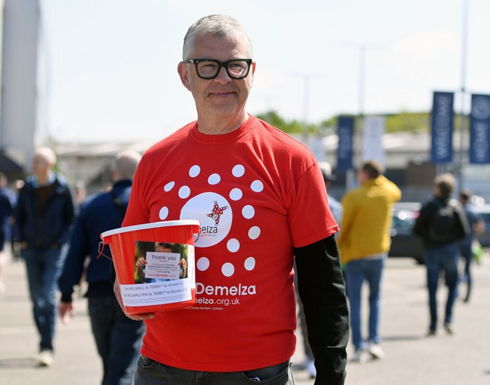 Sean is standing outside the Millwall Football Stadium, wearing a Demelza t-shirt, holding a collection bucket.
