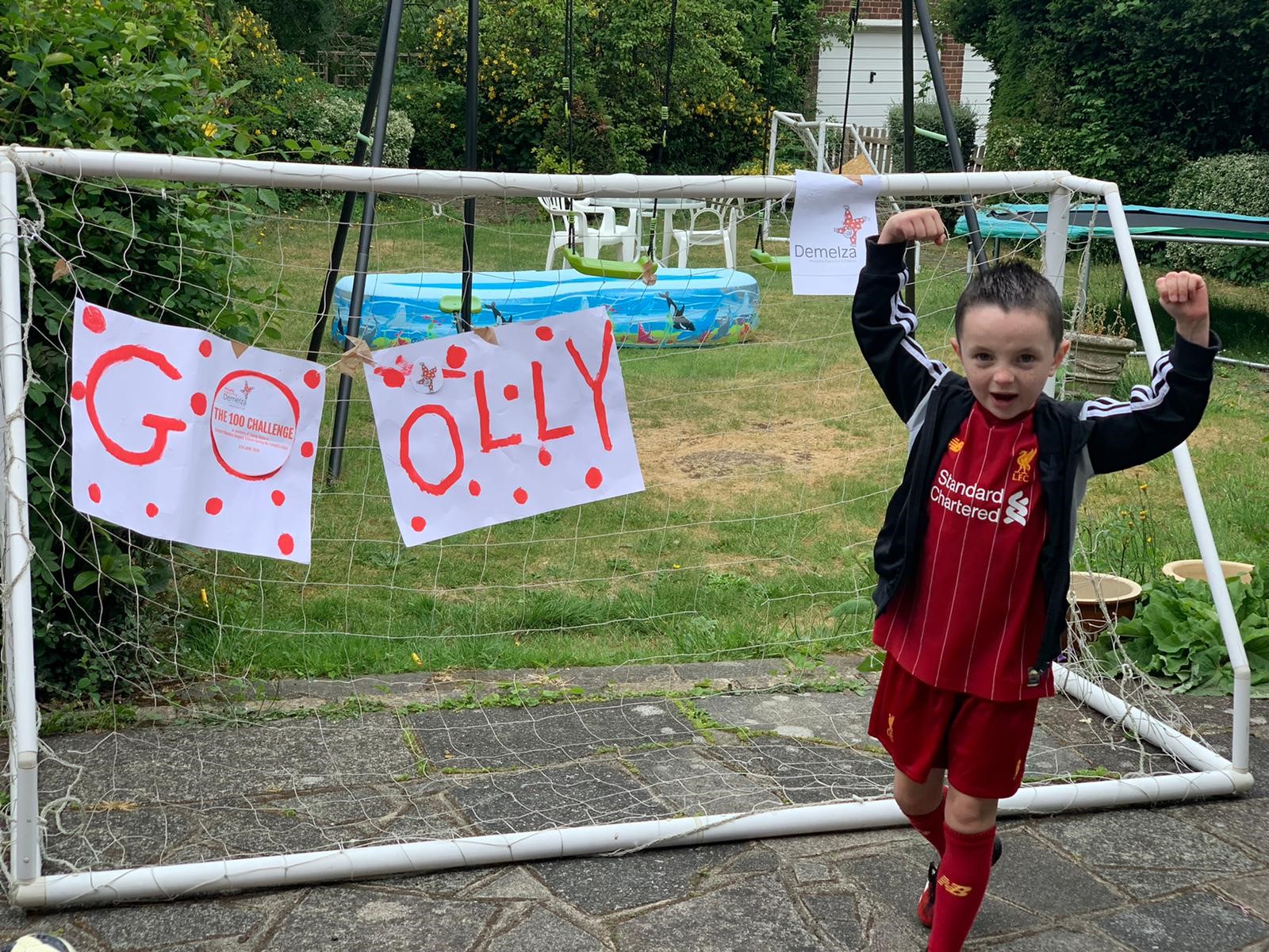 A boy celebrates completing his fundraising challenge. He is standing in front of a football goal with a 'GO OLLY' sign.
