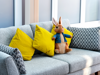 A grey sofa with two yellow pillows and two patterned grey pillows. A stuffed rabbit toy wearing a blue jacket sits in the centre of the sofa. Large windows with white frames are visible in the background.