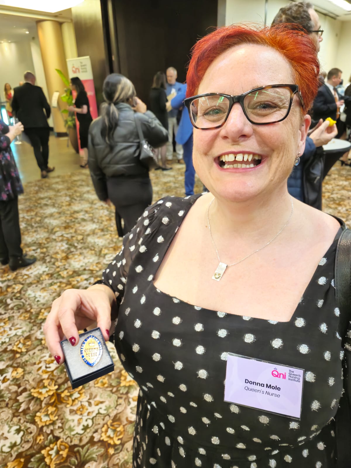 A person with red hair wearing a black dress with white polka dots holds an award in their left hand. They are standing in a room with a floral-patterned carpet and other people in the background, some engaged in conversation. A banner in the background reads "QNI." The person's name tag says "Donna Mole QNI Queens Nurse."