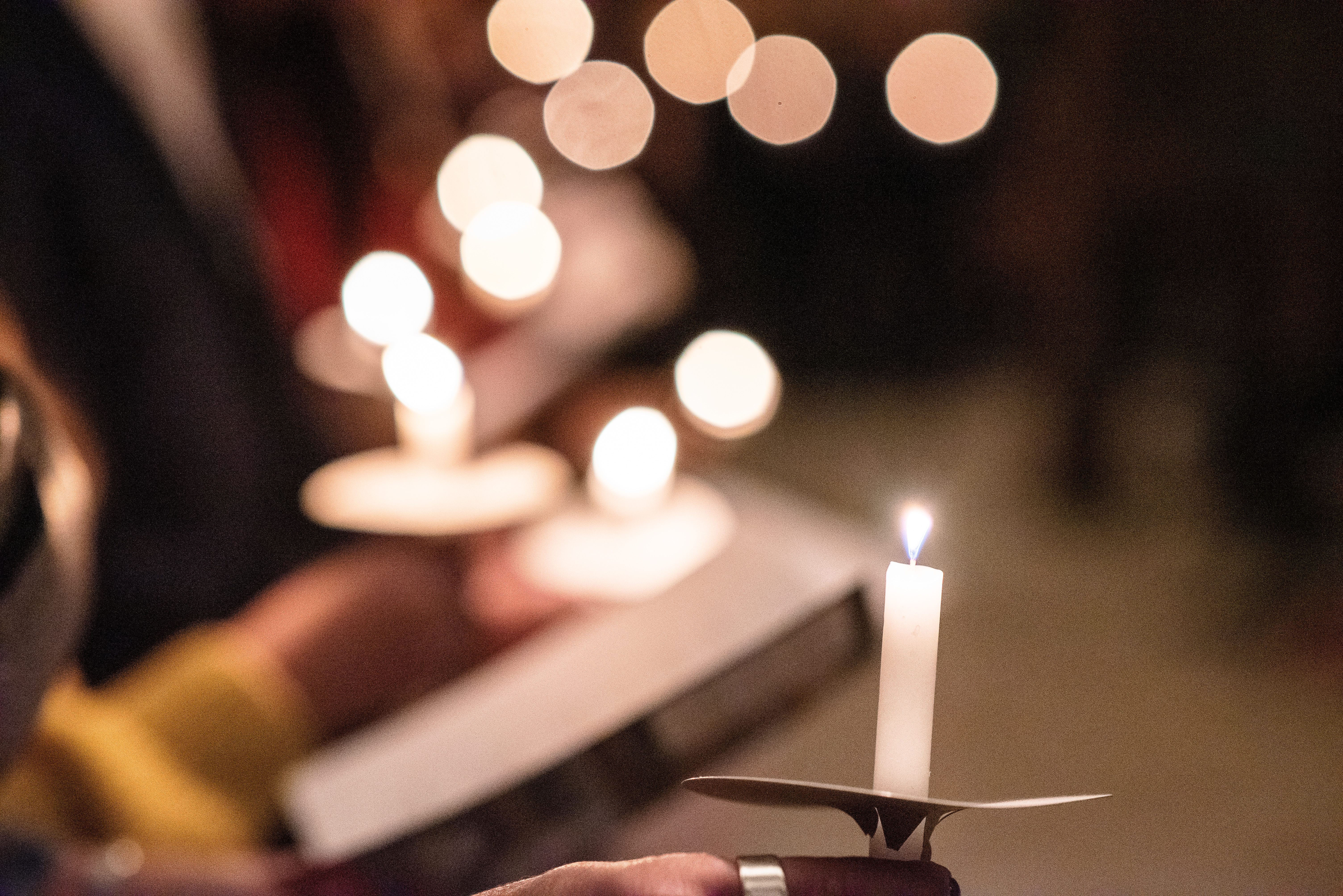 People holding lit candles in a dimly lit setting, with soft, blurred lights glowing in the background.