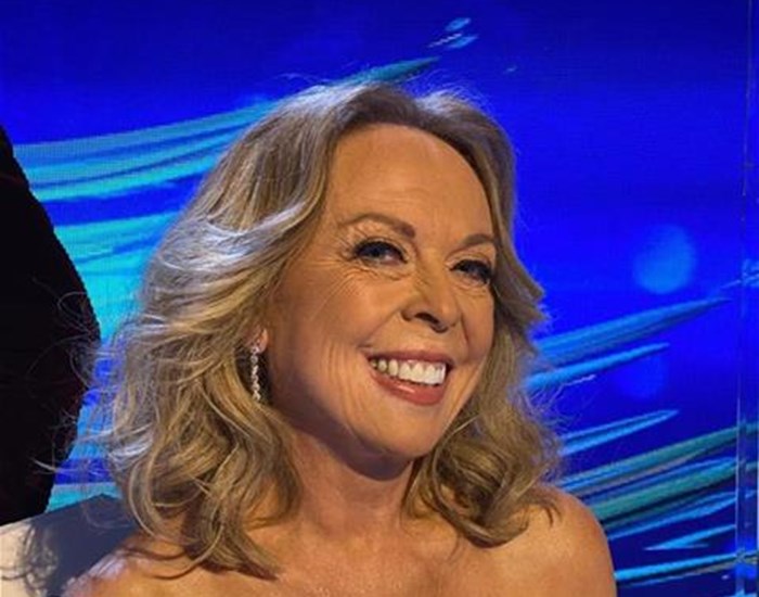 Jane Torvill wearing a glamourous black and white dress, sitting against a blue background, smiling. 