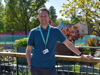 Man stands by a pond, wearing blue shirt, dark trousers, white shoes, and Demelza ID badge. Background includes trees, bushes, and a fence.