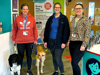 Three people stand indoors with two dogs. Behind them is a banner reading: 'Every day can be extraordinary. Give today and double your impact for children with serious conditions.'
