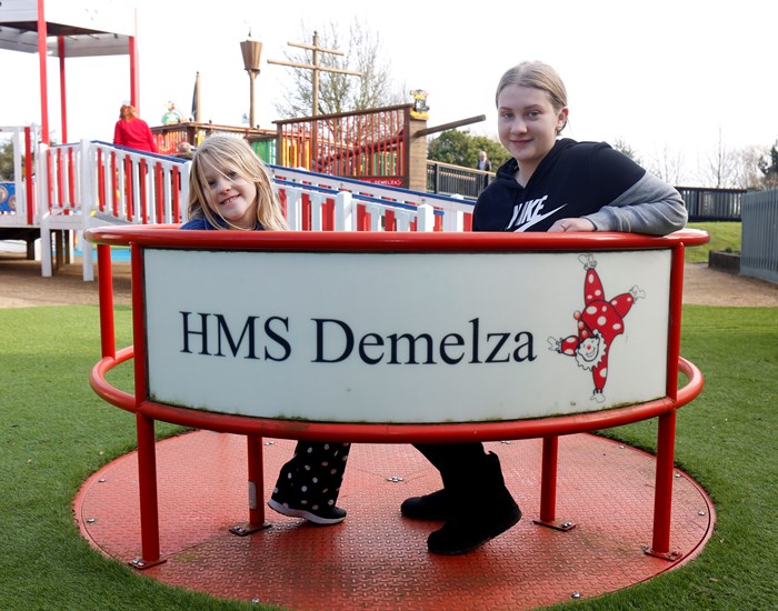 Two children sit on HMS Demelza, a round-about in the playground at Demelza Kent.