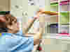 Nurse Becky in blue uniform reaches into a yellow-lidded bin labelled by Room number to sort medications in a storage unit.