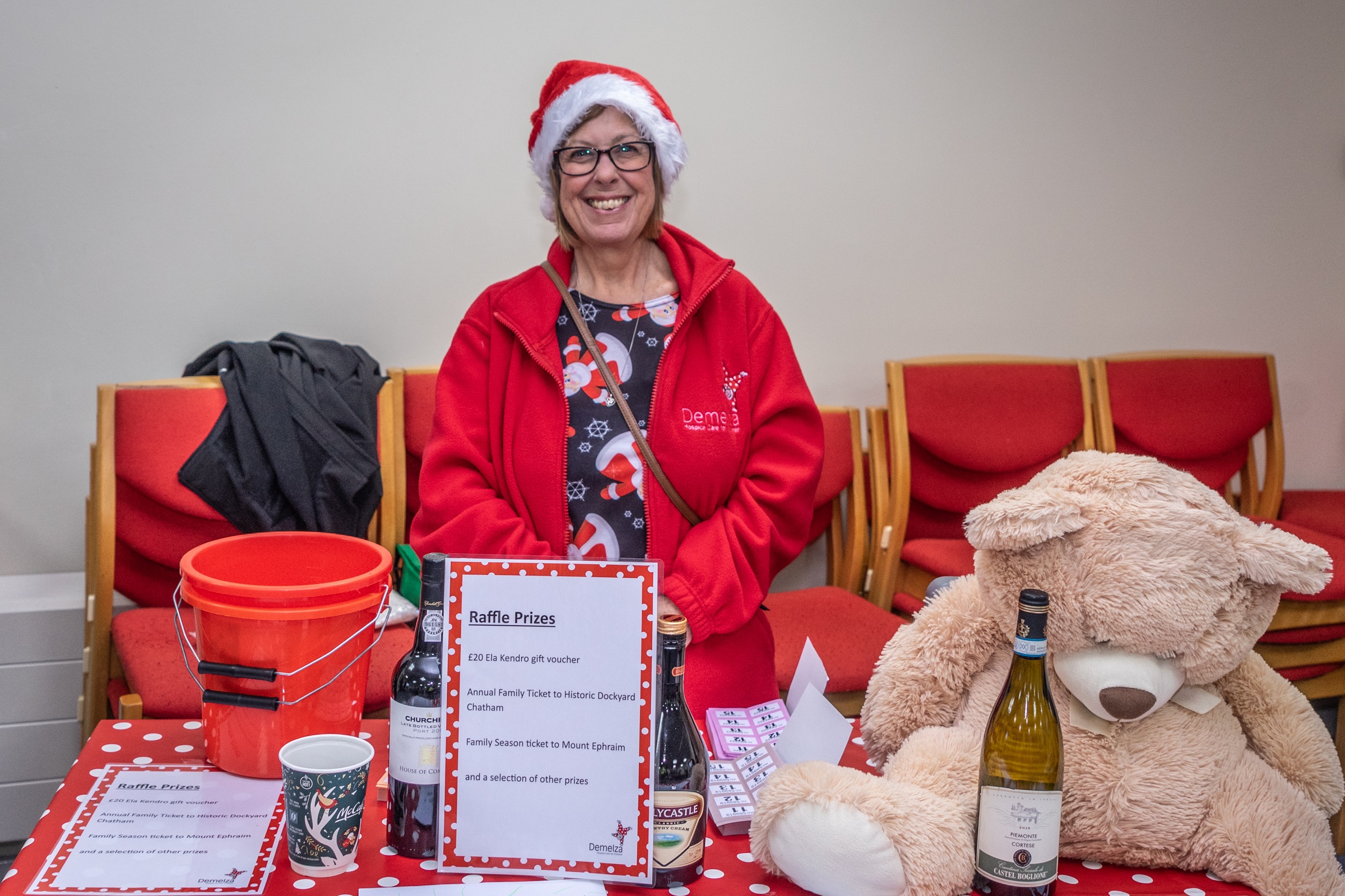 A woman stands behind her raffle table, smiling and wearing a Demelza fleece and Christmas hat.