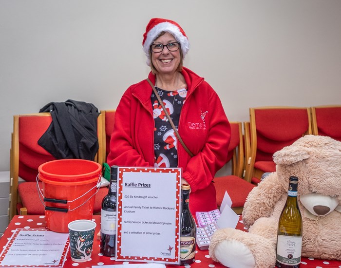 A woman stands behind her raffle table, smiling and wearing a Demelza fleece and Christmas hat.