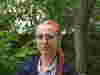 A person with long red braided hair stands outdoors in front of green foliage and trees. They are wearing a blue jacket over a light-colored shirt and have a Demelza identification badge hanging from their neck.