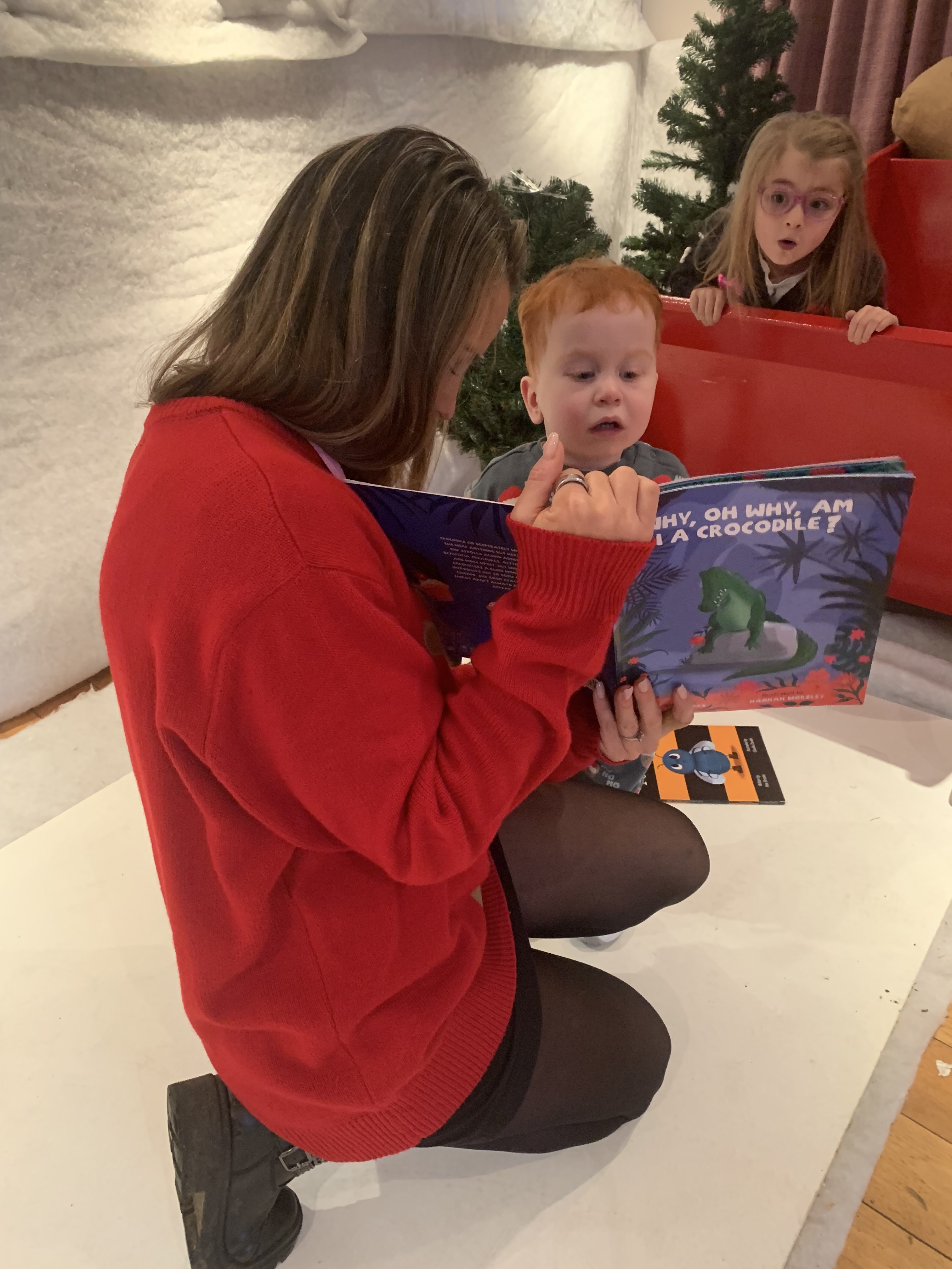 Person in red sweater kneels and reads 'WHY, OH WHY, AM I A CROCODILE?' to two children on a red chair.