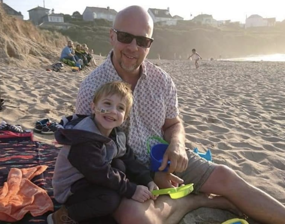 Zak and his dad sitting together on a sandy beach at sunset, smiling and holding buckets and spades.