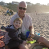 Zak and his dad sitting together on a sandy beach at sunset, smiling and holding buckets and spades.