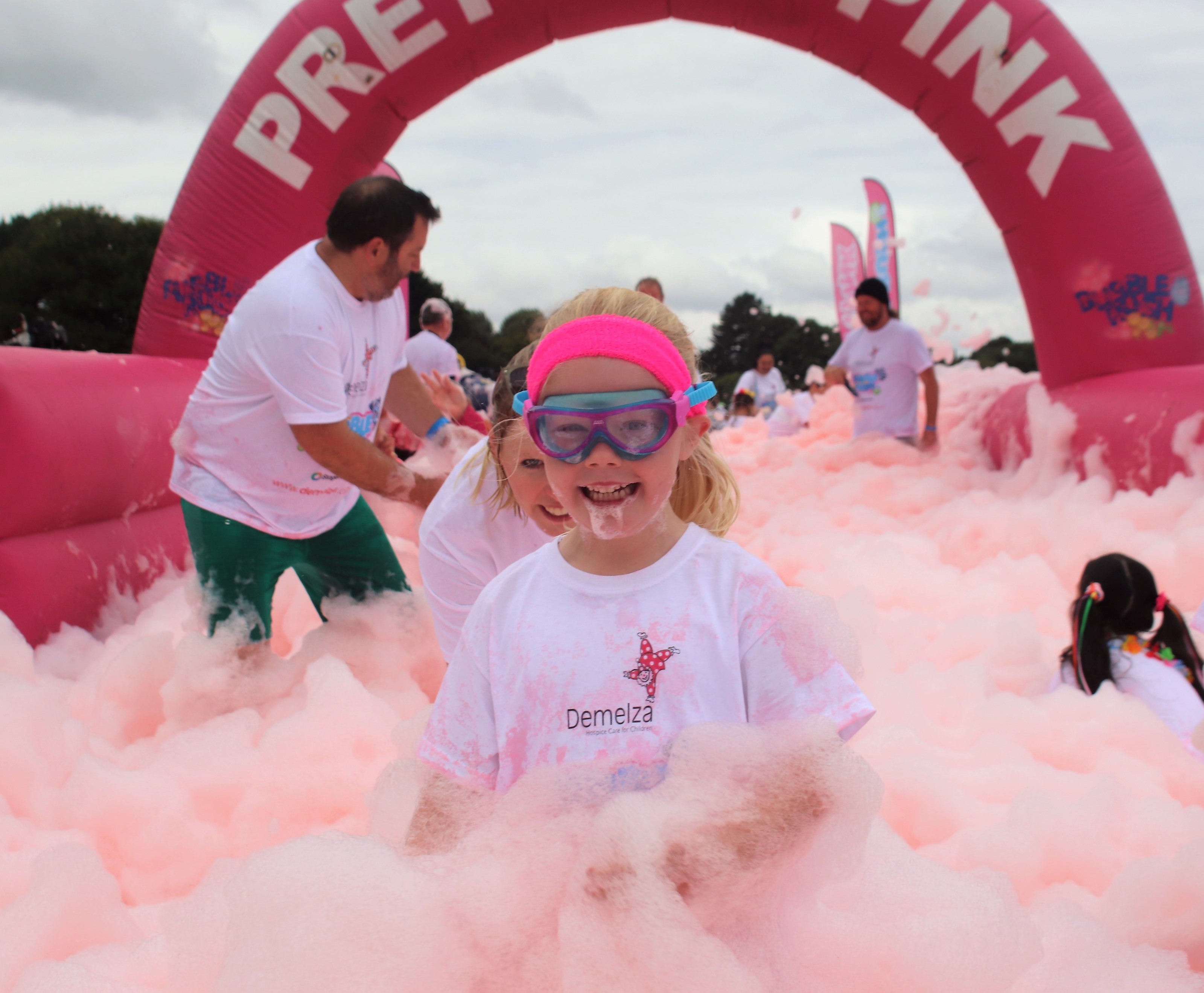 A young girl is standing in a sea of pink bubbles, wearing a pink headband and swimming goggles.